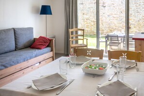 A dining area with a table set for two, a bowl of salad, and a view of the outdoors through large windows. - Appartement 300m de la Mer | Accès Piscine (Plougasnou)