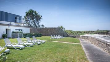 Une zone herbeuse avec des chaises longues blanches disposées en rangées, un mur de pierre et un bâtiment au design moderne.