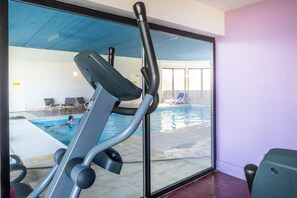 An indoor swimming pool with a view of the pool through a glass wall, a stationary exercise bike, and a few lounge chairs. - Appartement 300m de la Mer | Accès Piscine (Plougasnou)