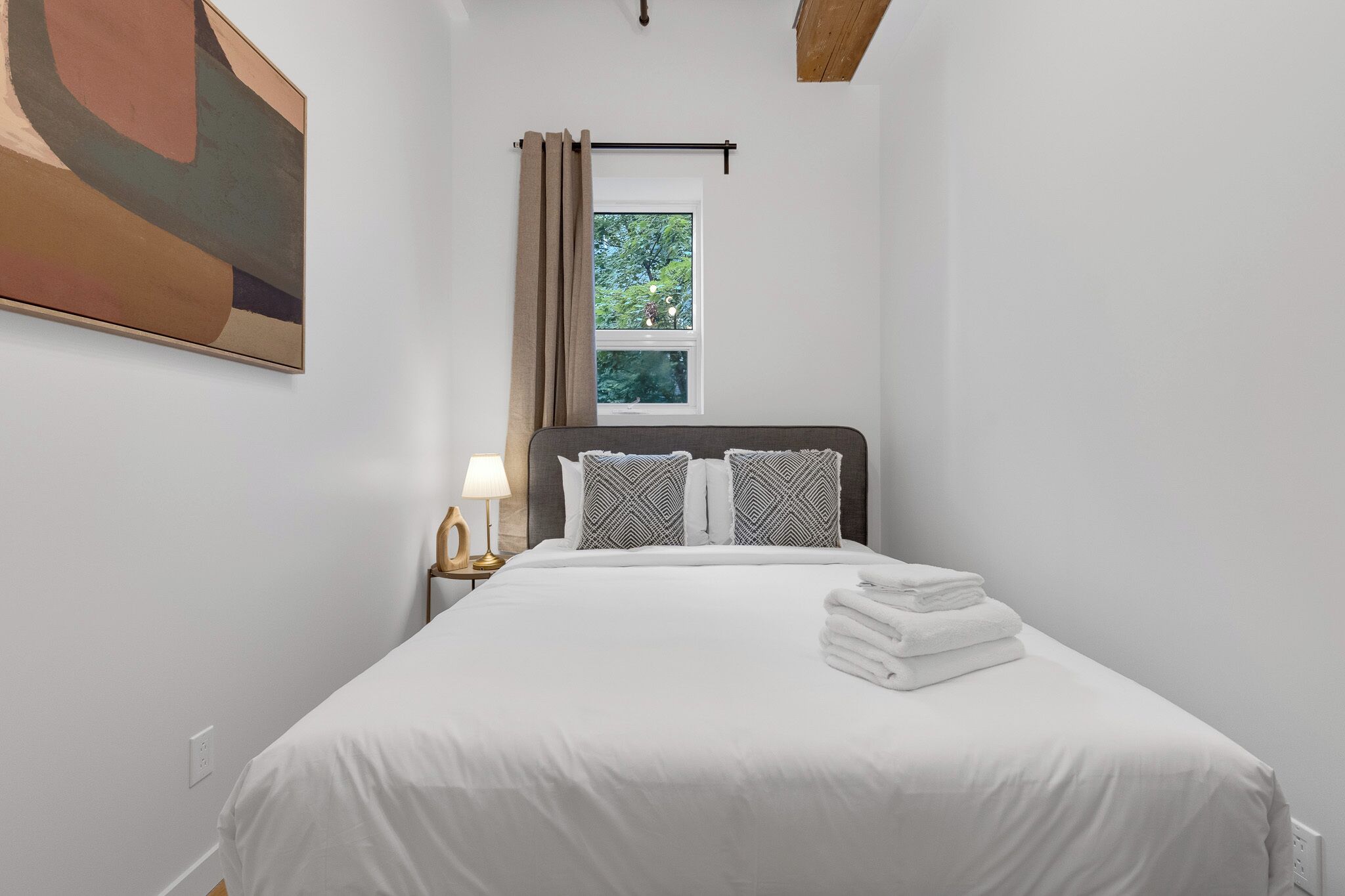 A neatly made bed with white bedding and two pillows, a window with curtains, a wall-mounted painting, and a bedside lamp.