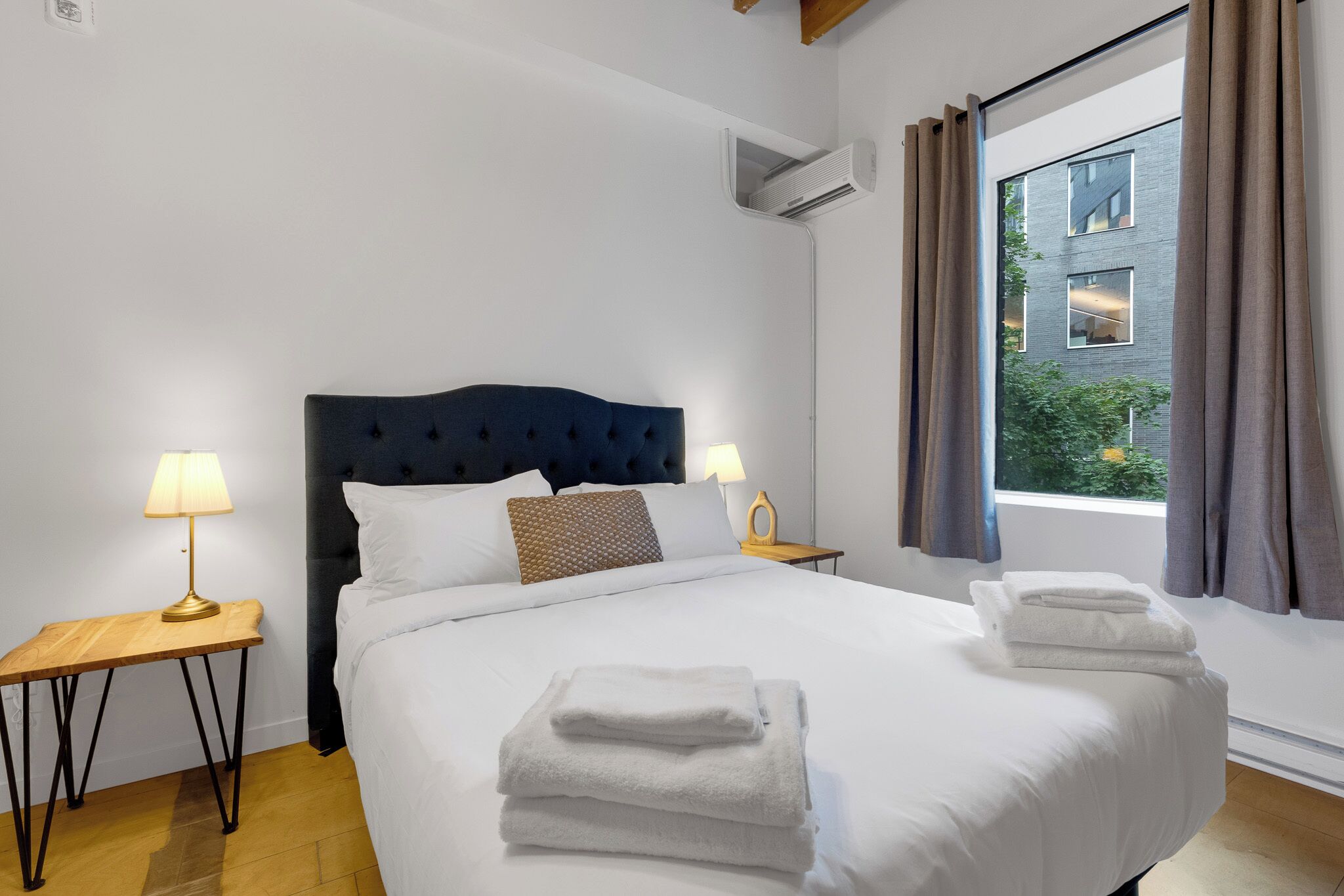 Cama bem arrumada com roupa de cama branca e toalhas dobradas, cabeceira estofada em azul escuro, duas mesas de cabeceira com abajures, e janela com cortinas.