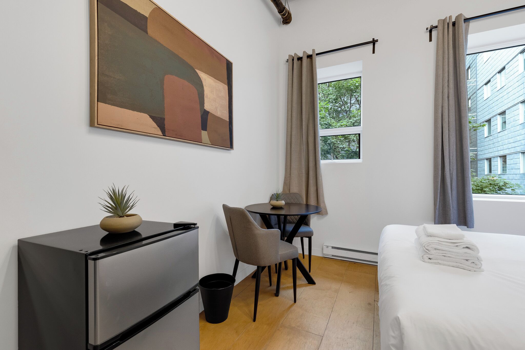A hotel room with a bed, a desk with a chair, a small refrigerator, a potted plant, and a painting on the wall.