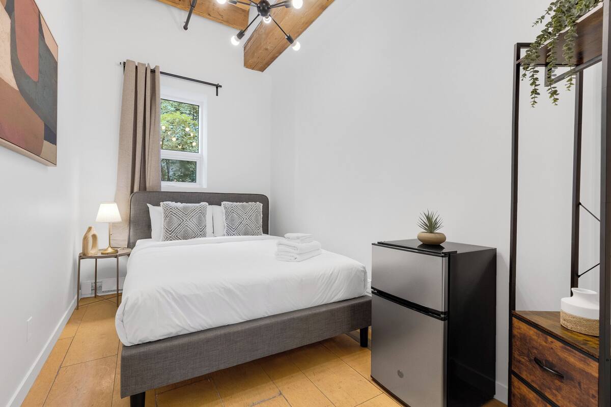 A bedroom with a bed, a nightstand, a refrigerator, and a window with curtains.