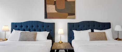 Habitación de hotel con dos camas, cada una con cabecera azul, ropa de cama blanca y una mesita de noche con lámpara.