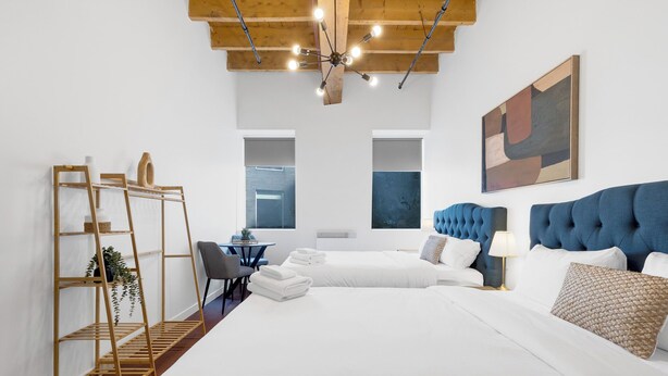 Une chambre d’hôtel moderne avec un grand lit, un bureau et une chaise. On y trouve une étagère avec des plantes, un tableau au mur et un luminaire au plafond.