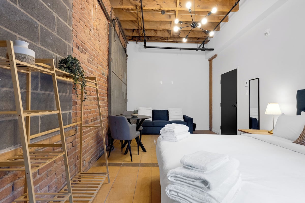 A hotel room with a bed, a desk, a chair, a small table, a sofa, a ladder, and a brick wall.