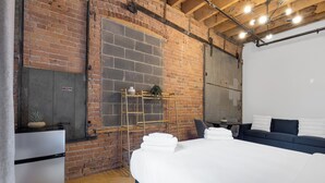 Une chambre d’hôtel moderne avec des murs en briques apparentes, un grand lit avec du linge de maison blanc, une petite kitchenette et un coin salon avec un canapé foncé et des coussins.