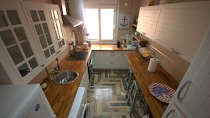 A compact kitchen with a wooden floor, white cabinets, a sink, a washing machine, and a window providing natural light. - 170 Tarek Puerto (Tarifa)