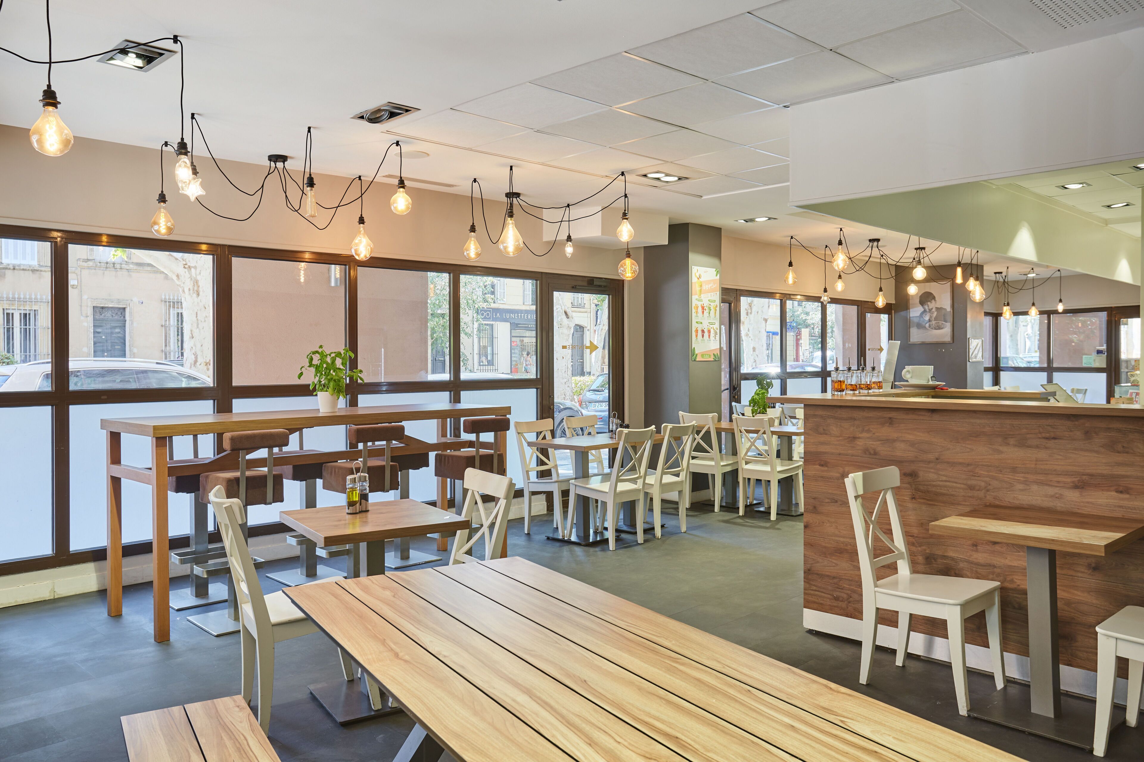 A modern cafe with wooden tables and chairs, large windows, and hanging light bulbs.
