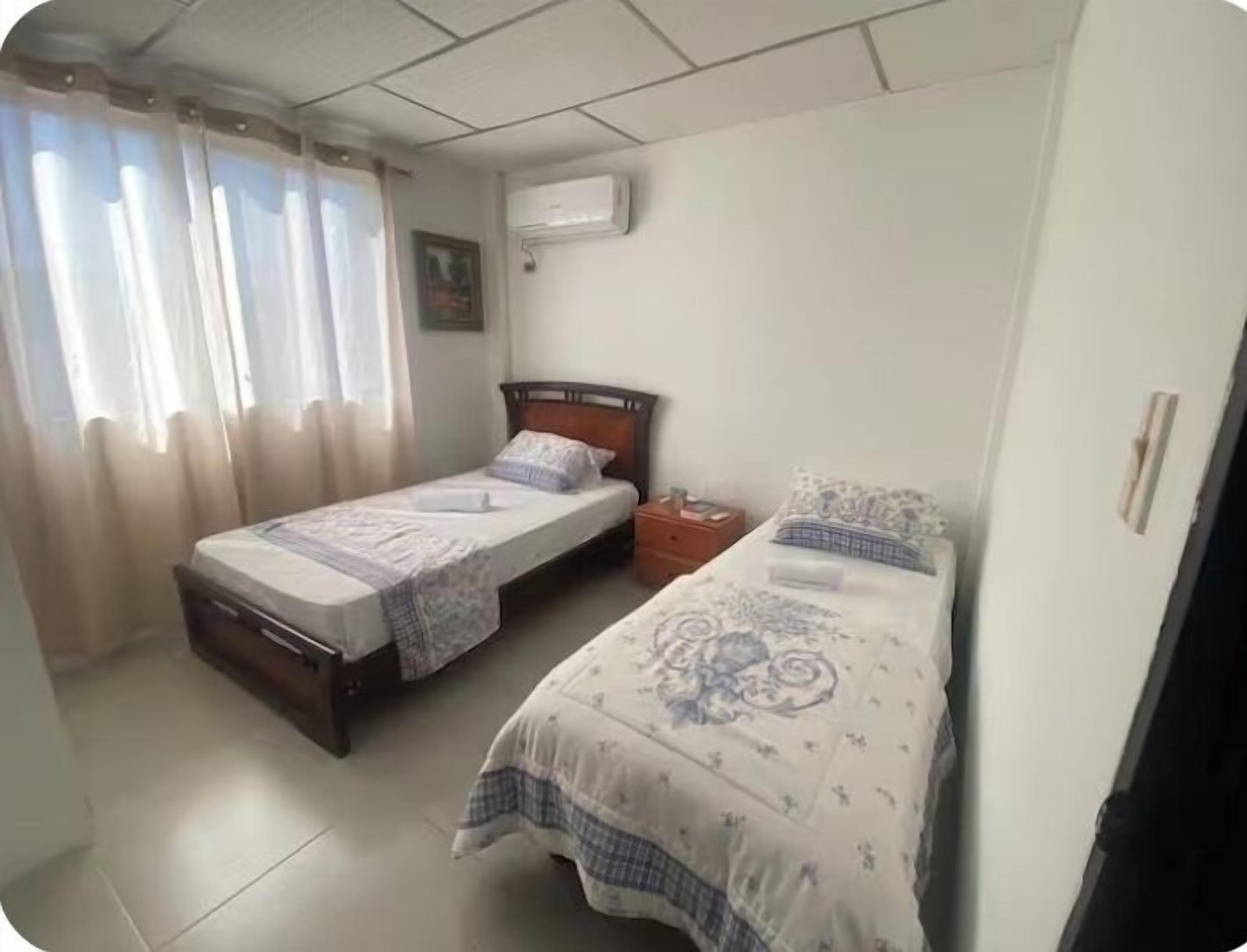 A small, clean room with two single beds, a nightstand, a window with curtains, and an air conditioning unit.