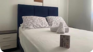 A neatly made bed with white bedding and floral pillows, a small bedside table with a drawer, a framed picture on the wall, and an air conditioning unit.