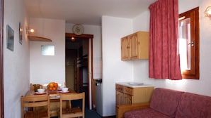 A compact room with a wooden dining table, chairs, a sofa, a small kitchenette, and a red curtain. - Studio abordable - Proche du funiculaire ! (Tignes)