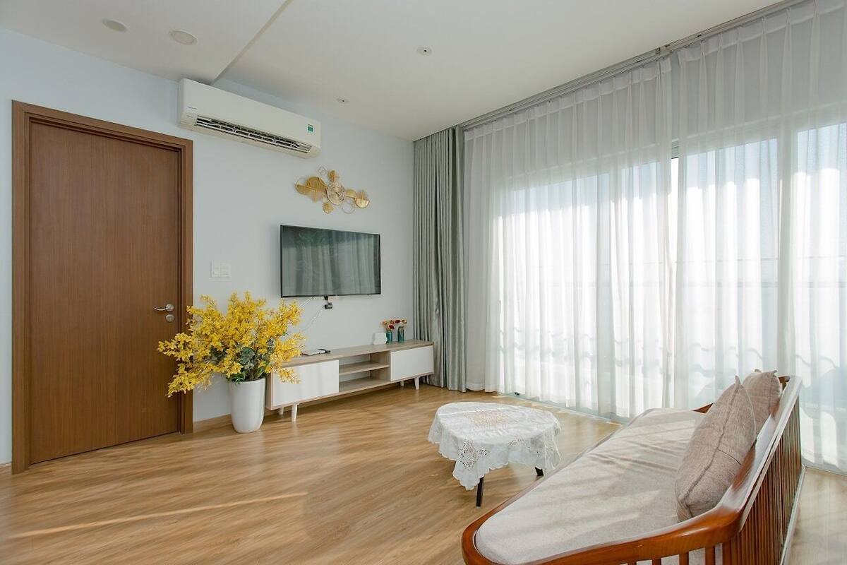 A modern living room with a wooden floor, a sofa, a TV mounted on the wall, a white console table, and a large window with sheer curtains.