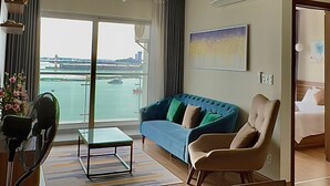 A modern living room with a blue sofa, a glass coffee table, and a dining area with a glass table. A large window offers a view of a body of water and a bridge. - Halo Bay Retreat (Hong Gai)