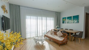 A modern living room with a sofa, dining table, and chairs, a TV, and large windows with curtains. - Halo Bay Retreat (Hong Gai)