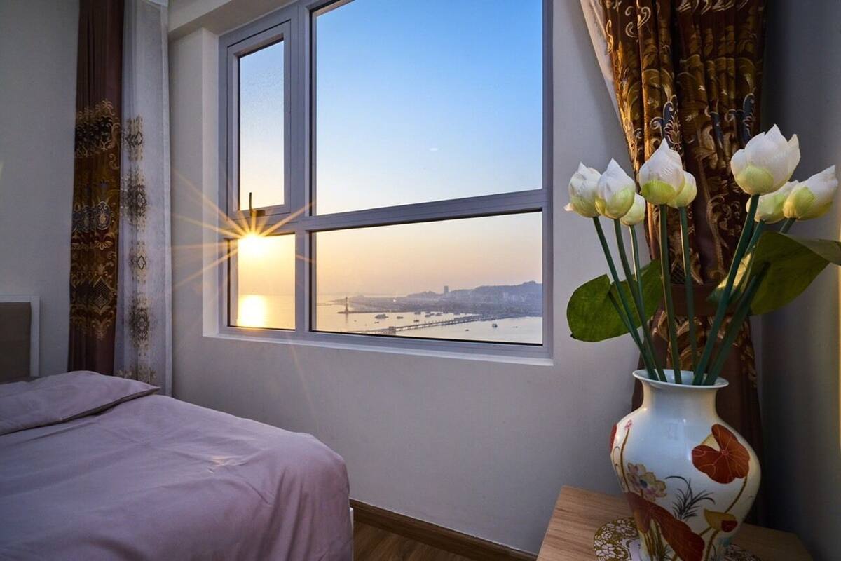 A bedroom with a large window, a bed, and a vase with flowers.