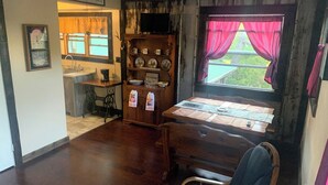 A cozy room with a wooden dining table, a blue chair, a wooden cabinet with dishes, and a window with red curtains.