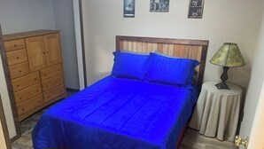 A bedroom with a wooden bed frame, a blue bedspread, a wooden dresser, and a small bedside table with a lamp.
