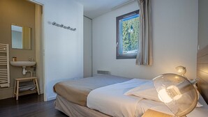 A bedroom with a bed, a window with a view of trees, a bedside lamp, and a bathroom with a sink and mirror. - Duplex 6p • Pied des pistes | Jacuzzi & piscine (FLAINE)