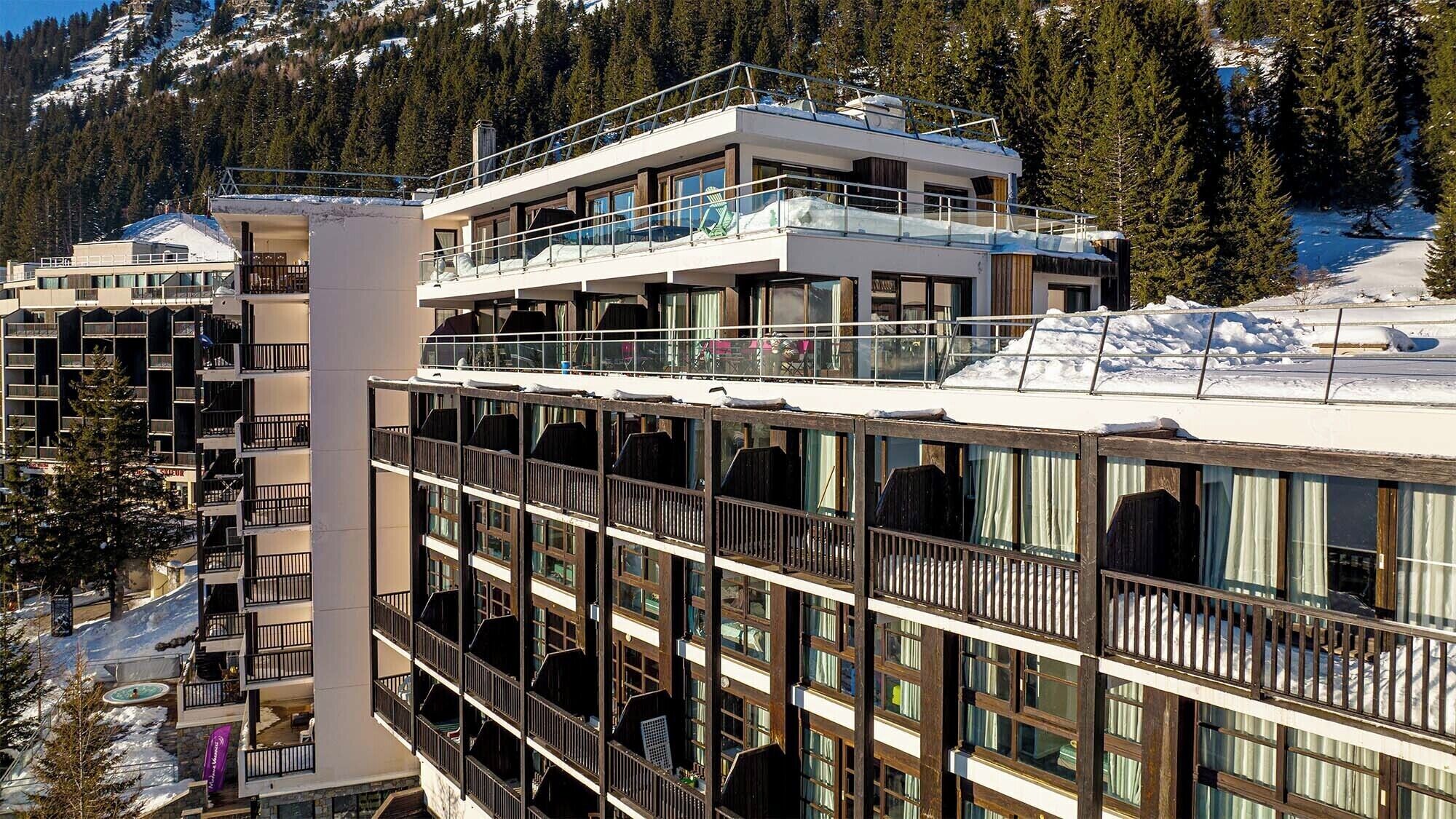 Un hotel de varios pisos con balcones, ventanas amplias y una montaña nevada al fondo.