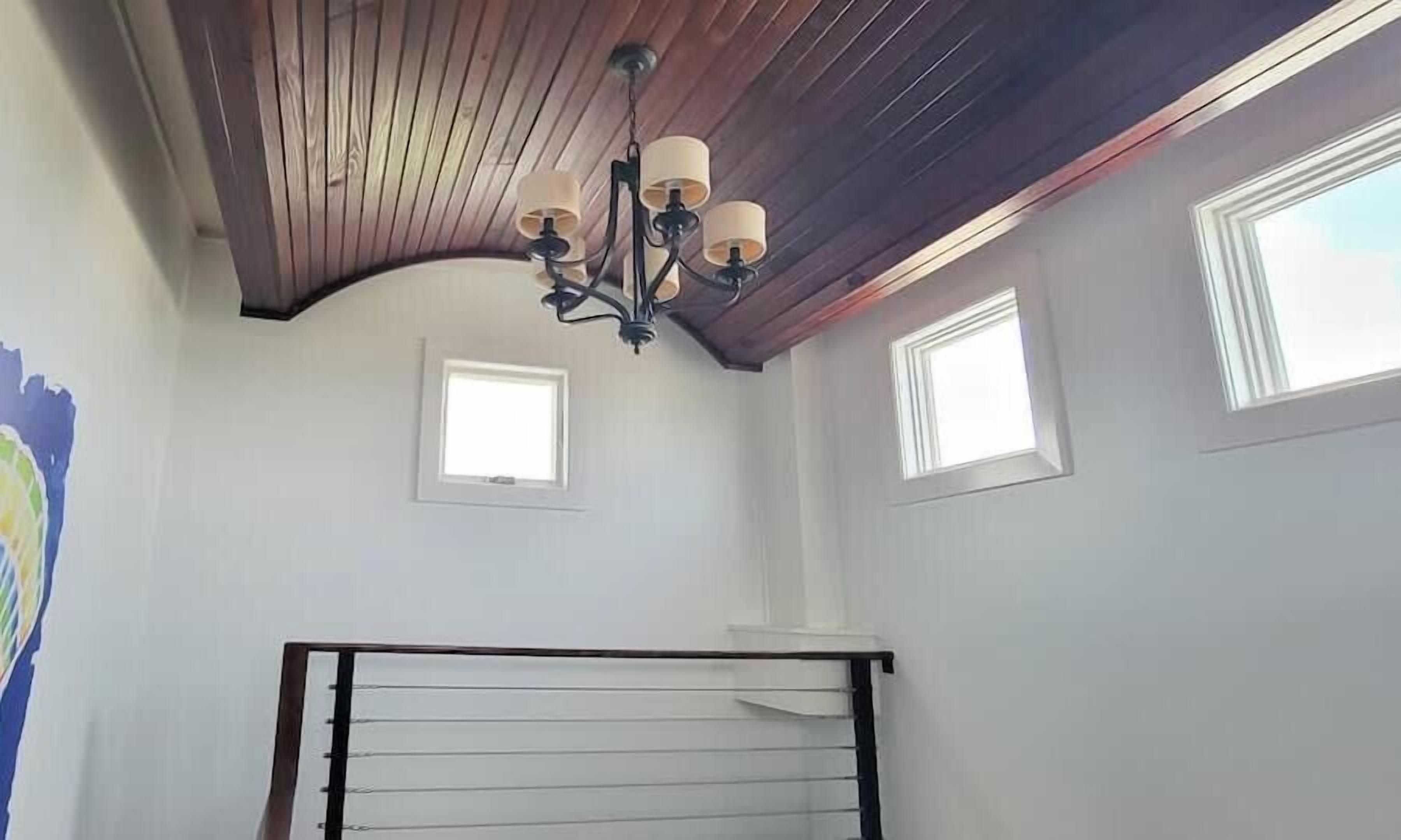 A room with a sloped ceiling, a chandelier, two small windows, and a bed with a wooden headboard.