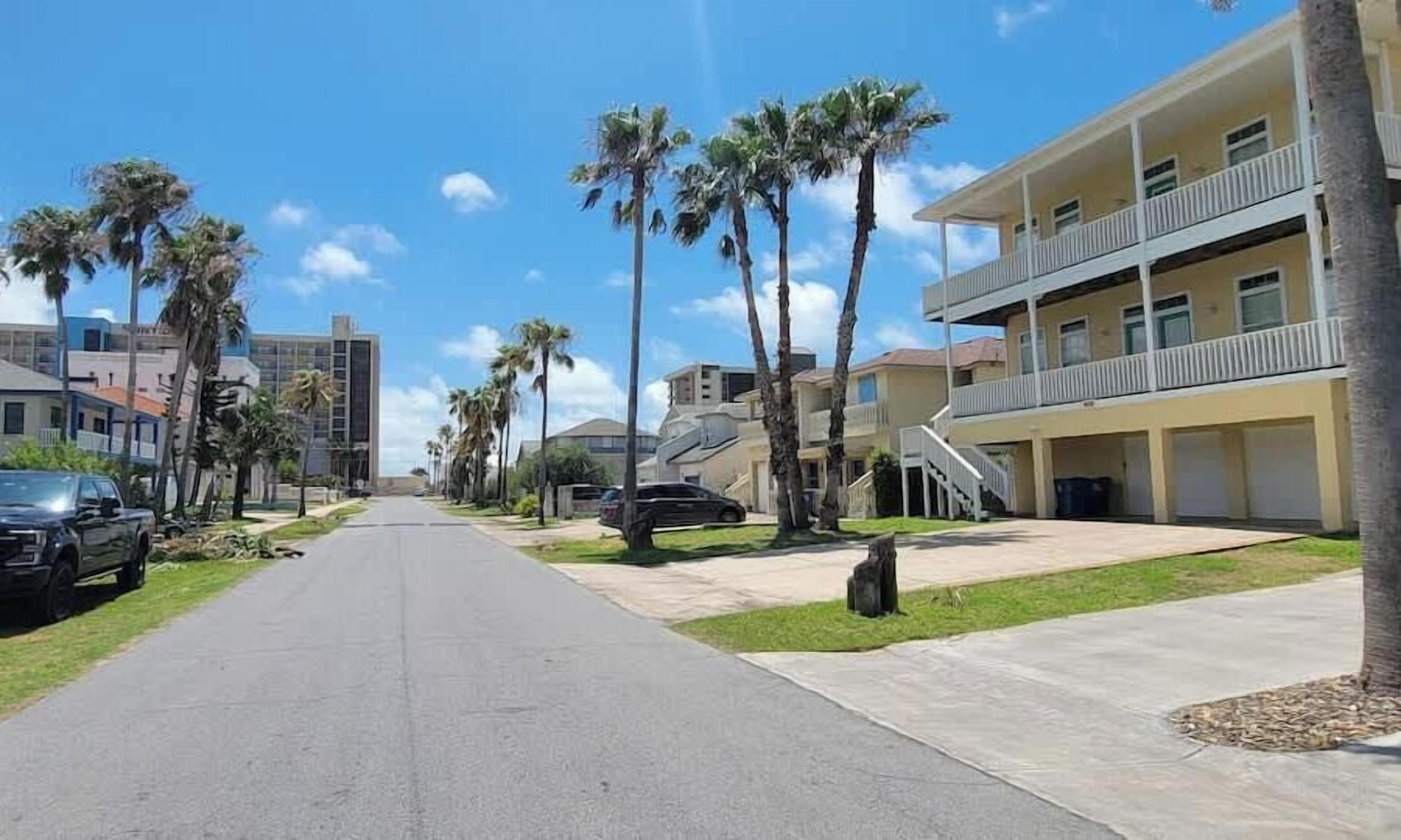 A residential street with palm trees, a multi-story building with balconies, and parked cars.