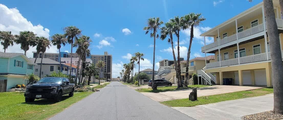 Una calle residencial con un edificio de dos pisos, palmeras y automóviles estacionados.