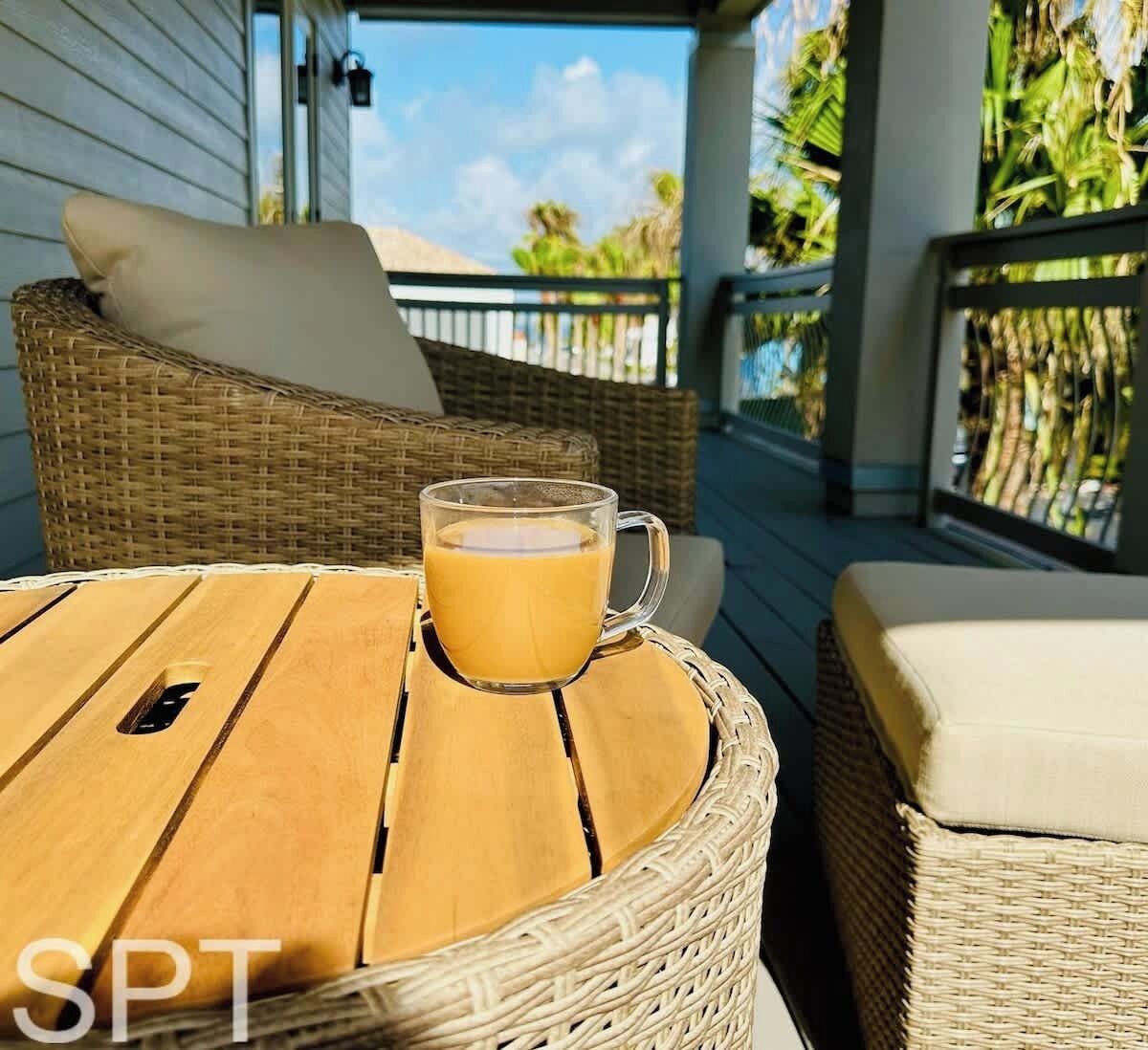 A wicker chair with a cushion, a wicker table with a glass mug of coffee, and a view of palm trees and a house in the background.