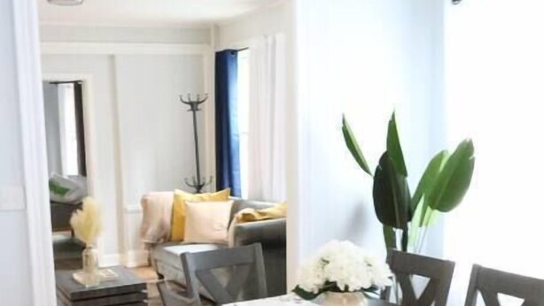 A dining area with a wooden table and chairs, a vase with white flowers, and a view into a living room with a sofa and plants. - 4 bedroom parking ,washer & dryer Apt (New York)