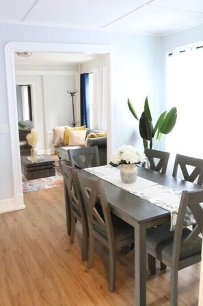 A dining area with a wooden table and chairs, a vase with white flowers, and a view into a living room with a sofa and plants. - 4 bedroom parking ,washer & dryer Apt (New York)