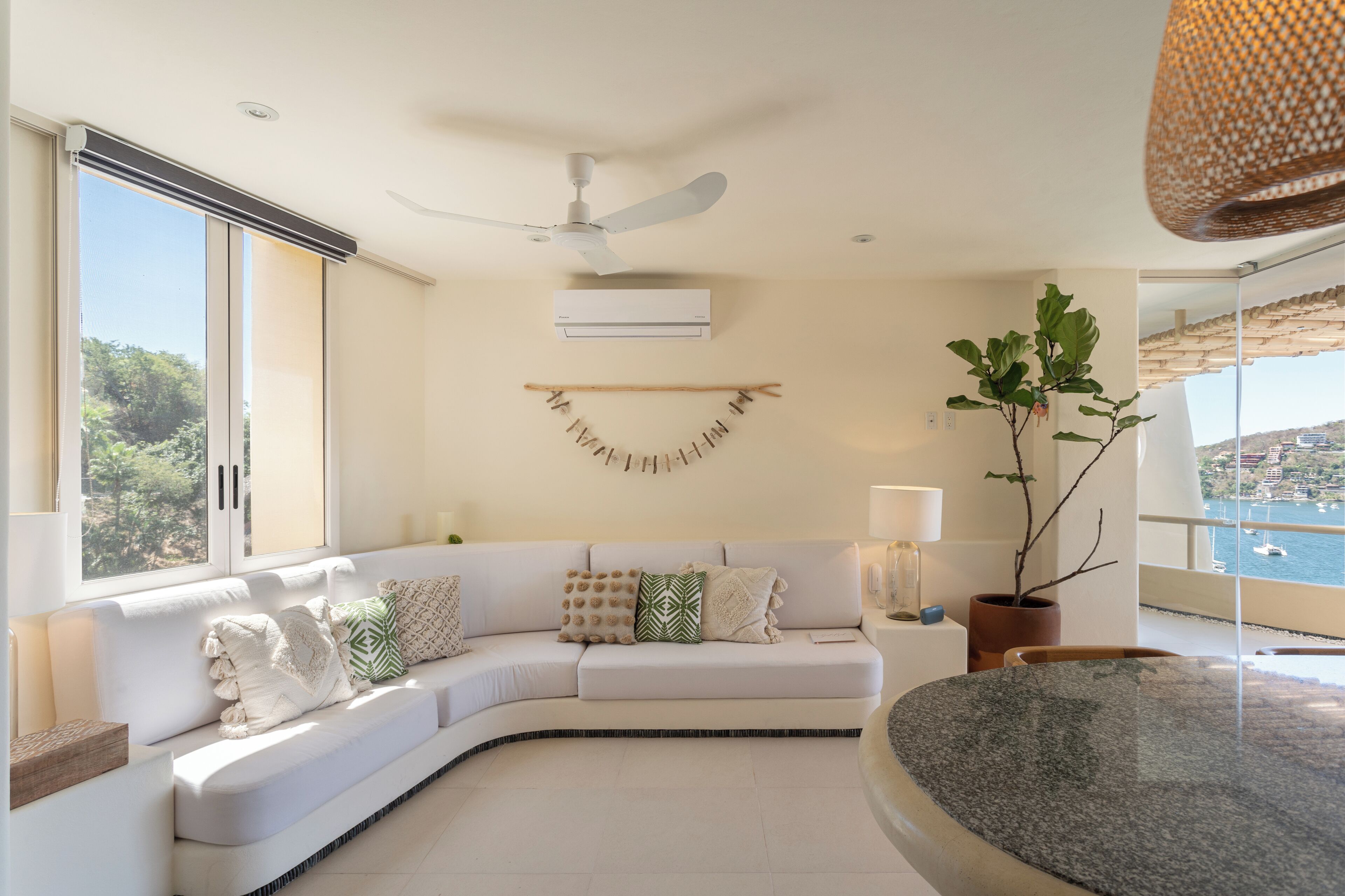 Een moderne woonkamer met een gebogen witte bank, een plafondventilator, een potplant en uitzicht op de zee door grote ramen.