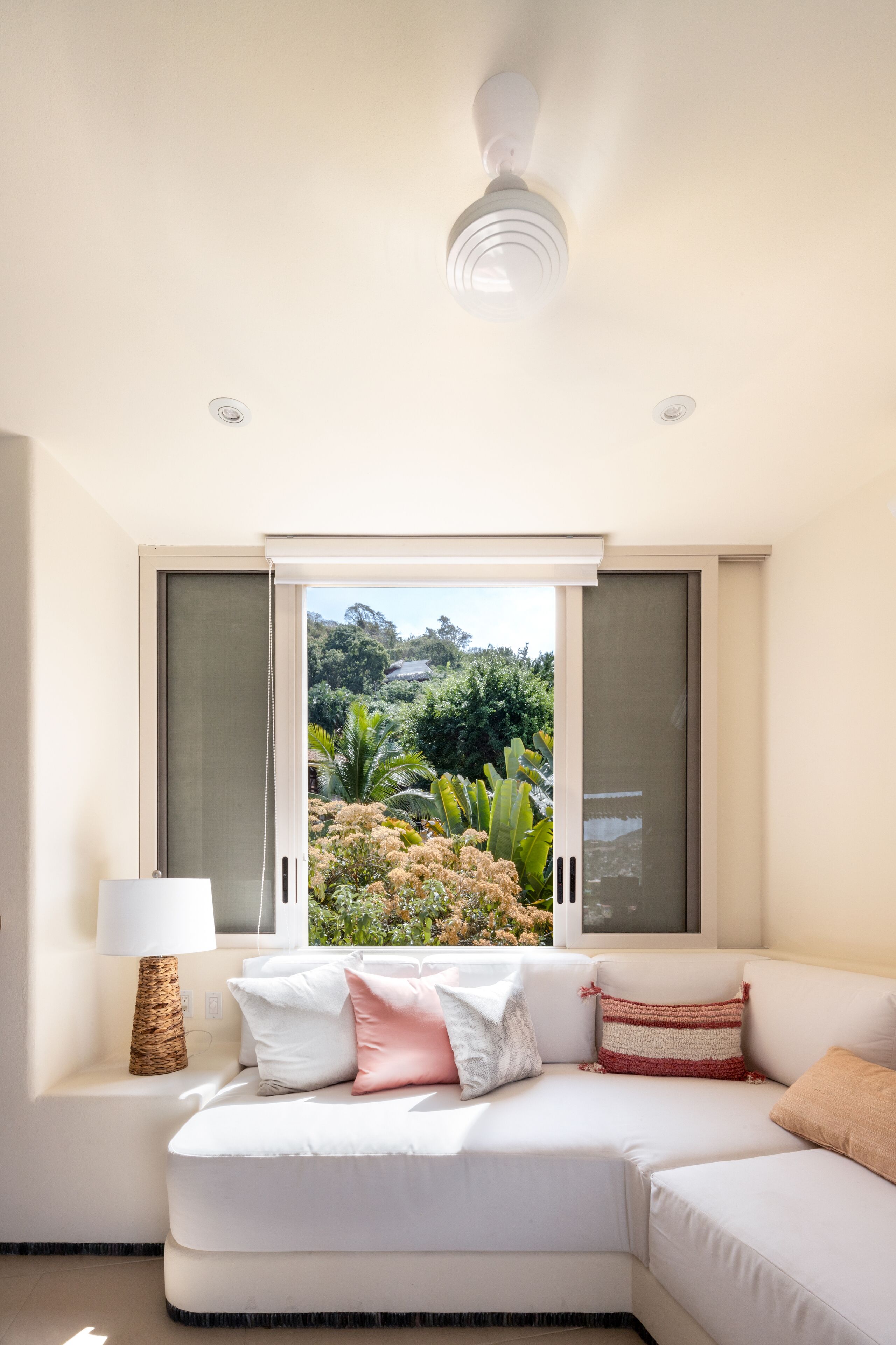 Un salon moderne avec un canapé blanc, une lampe et une vue sur la verdure par une fenêtre.