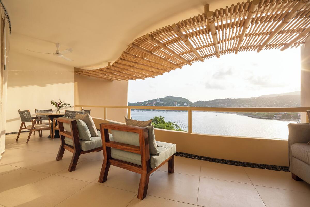 Un balcón con vistas a un lago y montañas, amueblado con sillas y una mesa de madera.