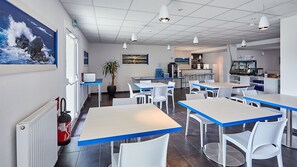 A modern dining area with white tables and blue-topped chairs, a kitchen in the background, and a large window with a painting of a seascape.
