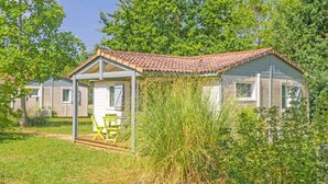 Une petite maison de plain-pied, avec un toit en tuiles, une véranda dotée d’une table et de chaises, le tout entouré de verdure et d’arbres.