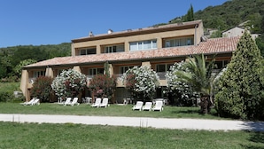 A two-story building with a tiled roof, multiple windows, and a balcony. There are several white lounge chairs arranged in front of the building. - Studio sur les Rives de l'Ardèche (VALLON-PONT-D’ARC)