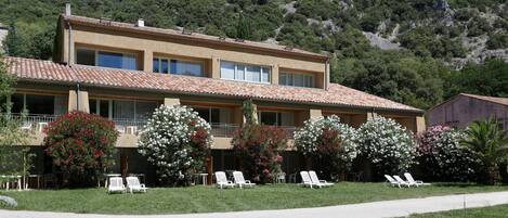 A two-story building with a tiled roof, balconies, and a garden with white lounge chairs.