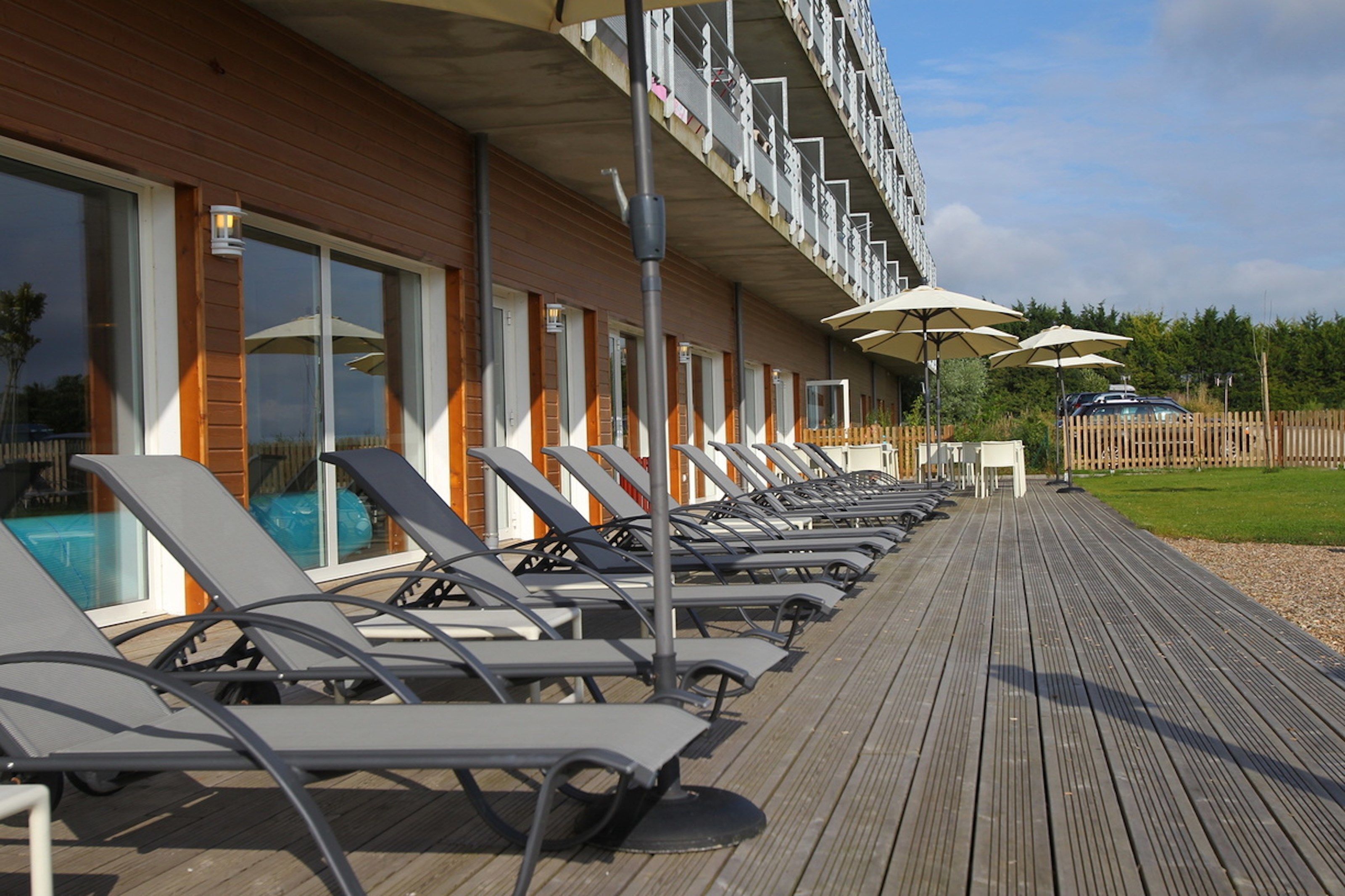 A poolside area with sun loungers and umbrellas, a wooden deck, and a building with large windows and a balcony.