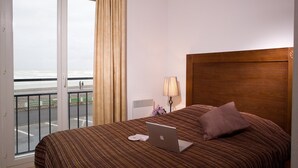 Una habitación de hotel con una cama, una laptop, una lámpara y vista a la playa a través de un ventanal grande.