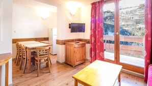 Una habitación con una mesa y sillas de madera, un televisor montado en la pared y un ventanal corredero que da a un balcón con vistas a un paisaje nevado.