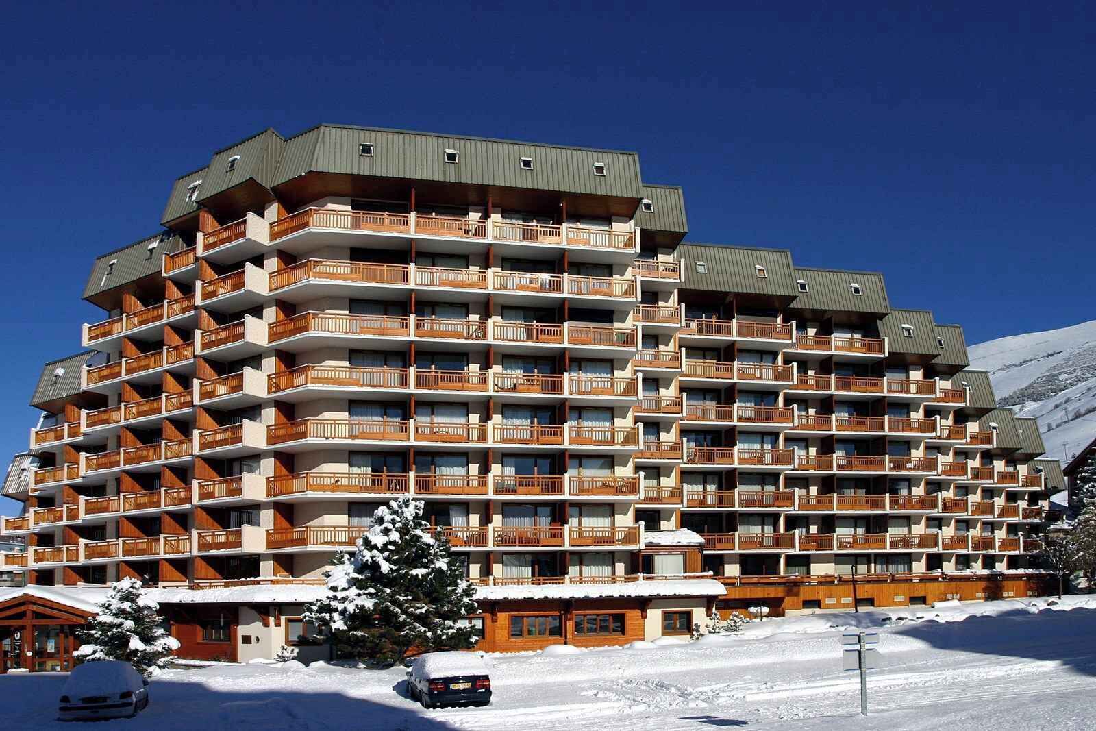 Um hotel com vários andares e varandas, com uma pista de esqui ao fundo e terreno coberto de neve.