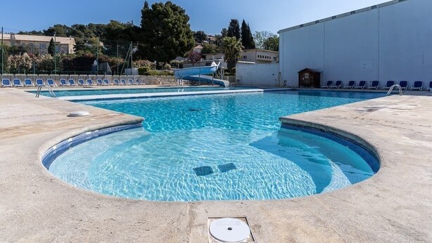 A swimming pool with a small slide, lounge chairs, and a building in the background.