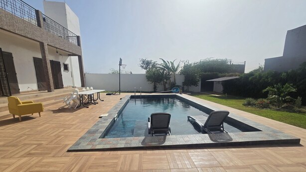 Une piscine entourée d’une terrasse en bois et d’un bâtiment blanc, avec des chaises longues et une table à proximité.