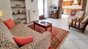 Una sala de estar con sofá, sillones, una mesa de centro y televisor. Se ve una cocina a través de un arco.
