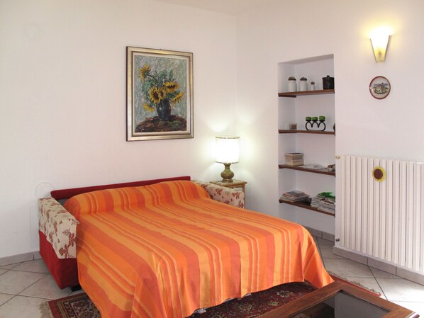 A bedroom with a bed, a nightstand, a lamp, a painting, shelves, and a radiator. - Casa Riboni by Interhome (Luino)
