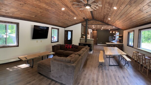 Un'ampia zona giorno con soffitto in legno, un divano grande, un tavolo da pranzo con panche e una cucina sullo sfondo.