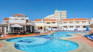 A hotel with a large outdoor swimming pool, multiple lounge areas, and a building with a tiled roof.