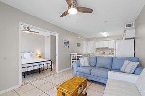 A living room with a sofa, a small table, a ceiling fan, and a kitchenette in the background.