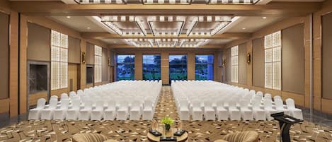 A conference room with rows of white chairs, a central table with a vase, and a view of greenery outside.