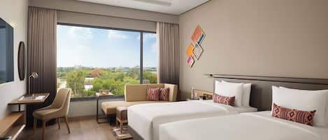Quarto de hotel com duas camas, uma escrivaninha, uma cadeira e vista para o verde.